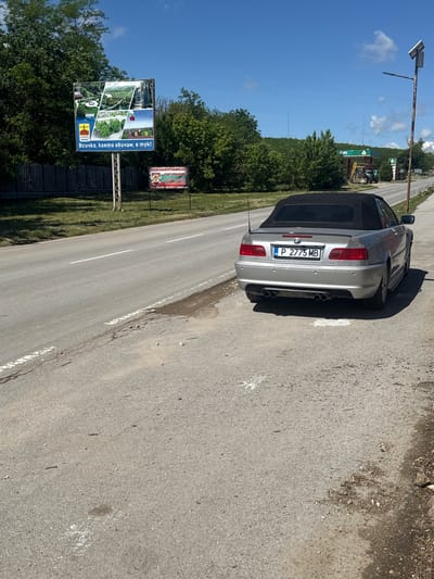 The image depicts a silver BMW convertible parked on the side of a road in Tsenovo, Bulgaria. The license plate number is P 2775 MB. Two billboards are visible on the other side of the road, one advertising "TEOBO" with the text "Всичко, което обичам, е тук!" (Everything I love is here!), and another advertising "БЕЗКОМПРОМИСНА СИГУРНОСТ" (Uncompromising Security) along with a phone number and website. There is also a grassy area with trees bordering the road. The sky is blue with some clouds. A lamp post is on the left.