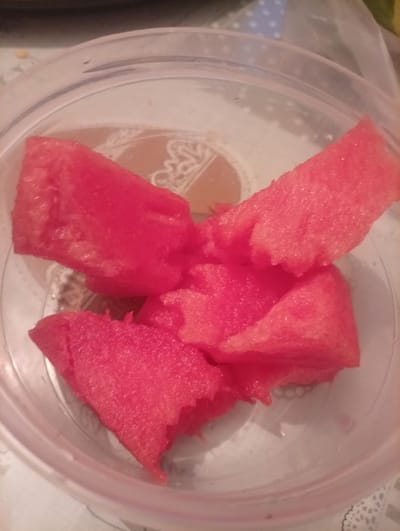 The image shows pieces of watermelon in a clear plastic container. The watermelon is a vibrant pink color and appears to be fresh. The container is clear and round, with some condensation on the inside. The background is blurred, but it appears to be a table with a patterned tablecloth. There are no people visible in the image. The lighting is artificial, suggesting the photo was taken indoors. The overall impression is one of a simple snack. The context of Shagonar, Russia, does not provide any specific visual cues within the image itself.