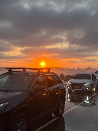 A black SUV with a roof rack is parked in the foreground, facing away from the viewer. The SUV is wet, indicating recent rain. To the right of the SUV, a white sedan is also parked, facing the viewer. The sedan's license plate reads "BWTL036". Both vehicles are parked on a wet asphalt surface, with white lines marking parking spots.

In the background, a pier stretches across the ocean. The sun is setting over the water, casting an orange glow on the clouds and the ocean. The sky is filled with dramatic, dark clouds that are beginning to break, allowing the sunset colors to shine through. There are waves crashing against a rocky shoreline between the cars and the pier. The image appears to have been taken in Oceanside, California, given the presence of the pier and the coastal environment.