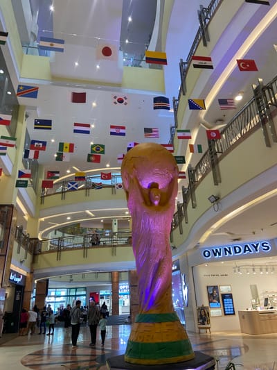 The image depicts a large shopping mall atrium in Pekanbaru, Indonesia, decorated with numerous national flags suspended from the ceiling. A prominent, large-scale replica of the FIFA World Cup trophy, illuminated with purple and green lights, stands in the foreground, slightly to the right of center. The atrium features multiple levels of balconies and railings, with shoppers visible on the lower levels and on a balcony in the background. Stores line the perimeter of the atrium, including "OWNDAYS" and other retail establishments with visible signage. The lighting suggests it is daytime, with bright overhead lights illuminating the indoor space. The overall atmosphere is commercial and festive, likely in anticipation of or celebration of a sporting event.