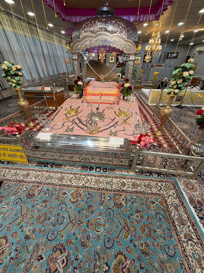 This image depicts the interior of a Sikh Gurdwara, likely in Chumkar, India. The scene is ornate and decorated with religious artifacts and floral arrangements. In the foreground, a large, intricately carved silver structure dominates the left side of the frame, possibly a decorative canopy or part of the shrine. It is adorned with purple and pink fabric drapes.
In the center of the image, a raised platform is covered with a pink and gold patterned rug. On this platform, several religious items are displayed, including what appear to be ceremonial swords and rings. A metallic railing surrounds the platform. Behind the platform, a large, light blue carpet with a detailed floral pattern covers a significant portion of the floor. Another patterned rug in shades of red and cream is visible underneath the platform.
Several bouquets of artificial flowers, in shades of white, pink, and red, are placed around the space, adding to the celebratory or reverent atmosphere. Decorative lighting strands with small yellow bulbs are strung across the ceiling, suggesting the time of day is likely evening or that the interior is well-lit.
A yellow sign with text in a foreign language is partially visible on the right side of the platform. The text appears to be "NOT ALLOWED" followed by inscriptions in Punjabi and Hindi. The overall impression is one of a sacred space, meticulously maintained and decorated for worship or a special occasion. There are no people clearly visible in the frame.