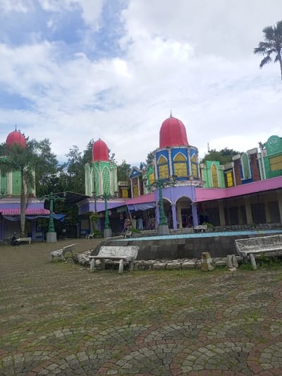This image depicts a wide exterior view of a colorful commercial or recreational complex in Cipanas, Indonesia, under a cloudy sky. The scene is dominated by a series of structures with vibrant, multi-colored facades, including prominent red, purple, pink, light green, blue, and yellow elements. Multiple towers with conical red roofs are visible, two with green bases on the left and center, and one with a blue base and yellow arched windows on the right.
In the foreground, a paved plaza of interlocking grey, red, and brown bricks is visible, with patches of green grass growing between the pavers. Several worn, white-painted concrete benches are scattered across this area. A circular, tiled fountain or pool structure, partially lined with blue tiles and appearing empty, is situated in the mid-ground.
The buildings feature various arched doorways and windows. Several storefronts or stalls are apparent, some with purple or blue awnings extended, displaying merchandise such as clothing on racks. Other sections appear closed or shuttered. Teal-colored ornate lampposts are positioned throughout the complex.
One individual is seated on a white bench on the far left of the image, facing away from the camera. Another individual is partially visible inside one of the open stalls further into the complex. The background shows dense green foliage and trees, including a palm tree on the far right. The sky is largely covered with scattered white and grey clouds.