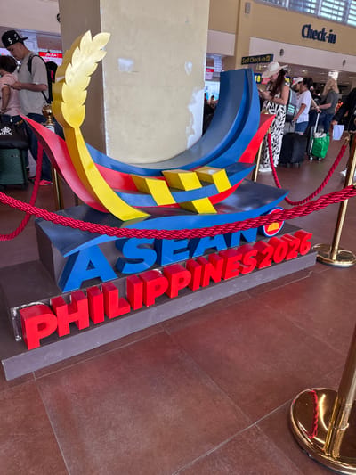The image depicts an interior scene at Bohol-Panglao International Airport in Panglao, Philippines. A prominent display for "ASEAN PHILIPPINES 2026" stands centrally, demarcated by red velvet ropes attached to brass stanchions. The display features a stylized boat-like structure in blue, red, and yellow, with a golden, leaf-shaped element on its left side. Below this structure, blue block letters spell "ASEAN", and red block letters spell "PHILIPPINES 2026". A small, circular ASEAN emblem is visible to the right of "ASEAN". The entire display rests on a grey, rectangular base.

In the background, several individuals are present, appearing to be airport passengers. On the left, a male wearing a black baseball cap and a light-colored t-shirt is observed looking at a mobile phone, with a black rolling suitcase nearby. A female is partially visible adjacent to him. On the right, a female wearing a wide-brimmed straw hat and a black-and-white patterned outfit is also looking at a mobile phone, standing near "Check-in" counters. Additional passengers and their luggage, including a black rolling suitcase and a green bag, are visible further in the background near "Self Check-in" signs. A light-colored concrete pillar with visible texture and patches stands behind the display. The floor throughout the scene is covered with large, reddish-brown square tiles.