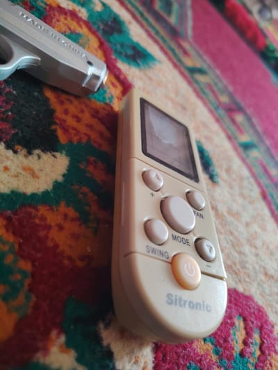 The image shows a close-up of an air conditioner remote control with the brand name "Sitronic" visible. The remote is light beige and has several buttons for functions like "SWING", "MODE", and "FAN". A small orange power button with a white power symbol is also visible. In the background, to the left of the remote, is a silver-colored toy gun with "MADE IN CHINA" stamped on it. Both objects are lying on a vibrantly patterned carpet with colors including red, orange, green, and black. The lighting suggests an indoor environment. There are no people or animals visible in the image. This scene is not identifiable to a specific location in Khujand, Tajikistan.
