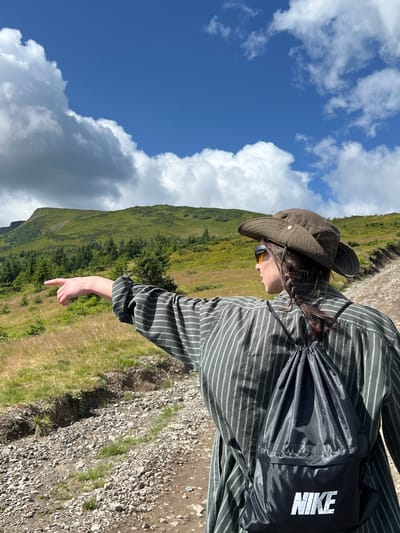 In the city of Chorna Tysa, Ukraine, a person wearing a striped shirt, sunglasses, a wide-brimmed hat, and a black Nike backpack stands facing away from the camera and points to the left. They are on a rocky, dirt path that winds up a grassy hillside dotted with evergreen trees. In the background, rolling green mountains rise under a bright blue sky with scattered white clouds. The lighting suggests it is daytime, likely afternoon given the angle of the sun.