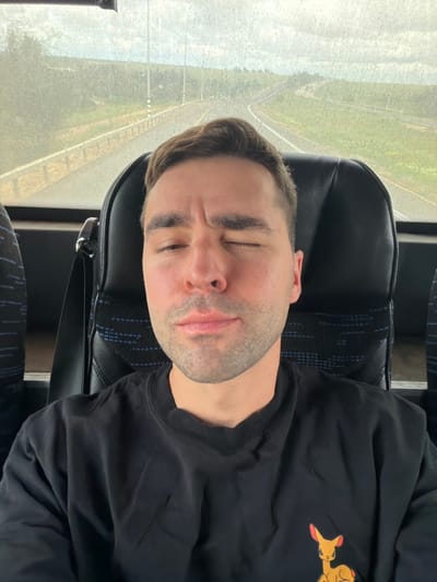 An adult male is depicted in a close-up, seated inside a bus in the Shafir Regional Council, Israel. He has short brown hair, light skin, and stubble, and is wearing a black long-sleeved shirt with an orange-eared cartoon deer emblem on the right chest. His right eye is closed, and his left eye is partially open, with a neutral mouth expression. A black seatbelt is visible over his left shoulder. The background, seen through a speckled window, shows a two-lane asphalt road with a metal guardrail, flanked by green grass and distant foliage under a cloudy sky. Several utility poles are visible along the roadside.