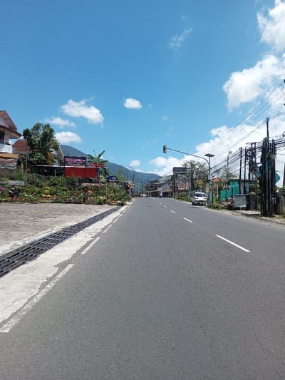 The image depicts a daytime outdoor scene of an asphalt road in the city of Bagochen Boolingpang Ueling, Bhutan. The sky is clear blue with scattered white cumulus clouds, illuminating the environment brightly. The road, which appears to be two lanes wide, features a dashed white center line and a solid white line marking its right edge. It extends into the distance, seemingly ascending towards a mountainous backdrop.
On the left side of the road, a concrete shoulder with dark metal drainage grates abuts an elevated embankment. This embankment is extensively planted with various flowering plants and green foliage. Behind the vegetation, multiple-story buildings are visible; these structures feature red-tiled roofs and facades predominantly in white or cream, with one building having a distinct red lower section and a dark sign displaying white text, possibly "MOONLIGHT."
The right side of the road begins with a dirt shoulder, transitioning into an area dense with utility poles supporting numerous tangled overhead electrical and communication wires. Further along this side, a streetlight equipped with a solar panel is present, along with additional buildings partially obscured by the utility infrastructure. In the mid-distance on the right side of the road, a white van is visible, either parked or moving slowly. A green traffic light is suspended over the road in the far distance, indicating an open intersection or passage. No people are discernible within the scene. The environment suggests a populated area integrated into a hilly or mountainous landscape.