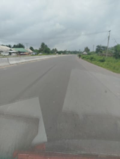 The media file shows an outdoor scene, likely on a cloudy day in Nigeria, possibly Odiderengwaji. A grey, paved road stretches into the distance, bordered by lush green vegetation on the right and a concrete barrier on the left. Buildings with white and green roofs are visible on the left side of the road, with a white van parked near them. A few figures, too indistinct to identify clearly, can be seen in the distance on the road, one appearing to be on a bicycle or motorcycle. The sky is overcast, suggesting a dim or potentially rainy day. The foreground is dominated by a blurry, grey surface, possibly a windshield or the hood of a vehicle, with a hint of an orange object beneath it. The overall impression is one of a journey along a rural or semi-rural road.
