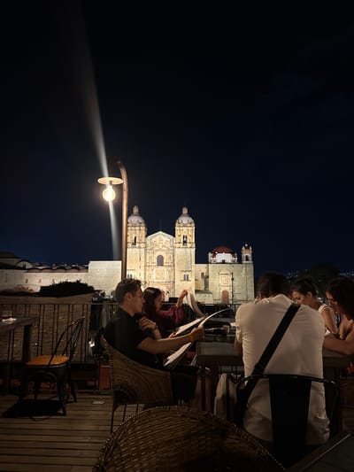 A low-angle, nighttime photograph shows a group of people dining at an outdoor restaurant in Oaxaca City, Mexico, with the Catedral de Nuestra Señora de la Asunción illuminated in the background. The perspective is oriented vertically. Patrons are visible in the upper and mid-sections of the image. They are seated, with several appearing to read menus. A man in a white shirt with a black strap across his shoulder is prominent in the upper right. Another person in a black shirt is in the center right.
In the mid-ground, a vintage-style street lamp is emitting a beam of light, which contrasts with the dark night sky. The cityscape, featuring the cathedral, is brightly lit.
The lower part of the image depicts the restaurant's outdoor setting, with wicker furniture and wooden decking. The architectural backdrop includes partial views of buildings adjacent to the restaurant and below the cathedral.