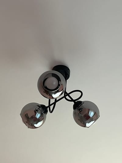 The image shows a close-up, low-angle shot of a modern, three-headed ceiling light fixture attached to a plain white ceiling. The fixture has a black central base from which three black metal arms extend. Each arm supports a chrome-finished, spherical lampshade with an opening at the top revealing a white light bulb. The reflective surfaces of the lampshades show distorted reflections of the surrounding environment, including parts of what appear to be other rooms or objects. The image is taken indoors, and there are no people or animals visible. The lighting is a mix of ambient light and the light from the fixture itself. The location is identified as Branipole, Bulgaria.