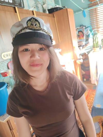 The image shows a young woman wearing a white captain's hat with gold trim and an anchor emblem, and a brown t-shirt. She is positioned in the foreground, looking directly at the camera. Her hair is brown and falls past her shoulders. The background appears to be an indoor domestic setting, possibly a kitchen or living area, with wooden cabinets and blinds visible. There are also some plants, containers, and a blue bucket in the background. The lighting suggests it is daytime, with sunlight streaming in from the right. The overall impression is a casual selfie taken in a residential environment. No specific location in Molodegniy, Russia is identifiable from the image.