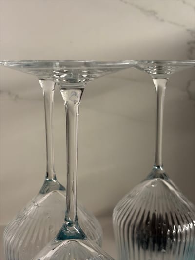 The image shows a close-up, low-angle view of three clear glass wine glasses, inverted and stacked. The glasses have ribbed bowls and slender stems. The background is a light-colored, textured surface, possibly a countertop or wall. The image appears to be taken indoors. The location of Holon, Israel, is not discernible from the visual information provided in the image.