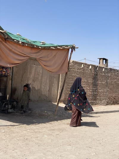 A woman in a floral burqa walks along a sunlit dirt road. A young child in olive clothing stands in the shadow of a makeshift dwelling, covered with a draped canopy. Behind them is a crumbling brick wall with an old tower. The clear sky is above.