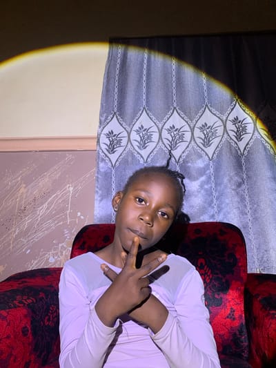 A young girl is seated on a red patterned armchair. She is wearing a light purple, long-sleeved top. Her hair is styled in small braids, with some strands pulled back from her face. She is looking at the camera and holding up two fingers in a peace sign. The armchair is decorated with a black floral pattern. Behind her, a grey curtain with a leaf pattern is visible. A spotlight illuminates the scene from the left side of the image, casting a warm glow on the curtain and the girl. The background wall appears to be painted a light color with some splatters of white and black paint. The overall lighting suggests it could be indoors at night, possibly with a portable light source or staged lighting. The girl's expression is neutral, but her gesture is friendly. The context provided places the location as Barakin Sabawa, Nigeria.