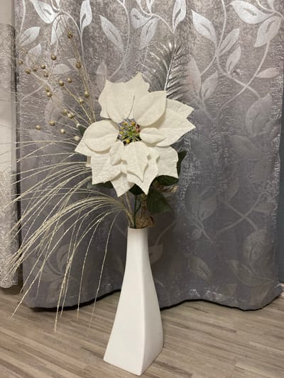 An artificial white poinsettia arrangement is displayed in a white vase on a wooden floor, with a silver-leaf patterned curtain in the background. The arrangement includes the poinsettia, along with decorative branches adorned with small gold beads and glittery, grass-like strands. The poinsettia's center has a colorful, glittery embellishment. The scene appears to be an indoor setting, likely a home or a shop, given the decorative nature of the objects and the presence of curtains and flooring typical of such environments. There is no indication of a specific location in Sachsenheim, Germany, as the image lacks any identifying landmarks or contextual clues to pinpoint a geographical location.