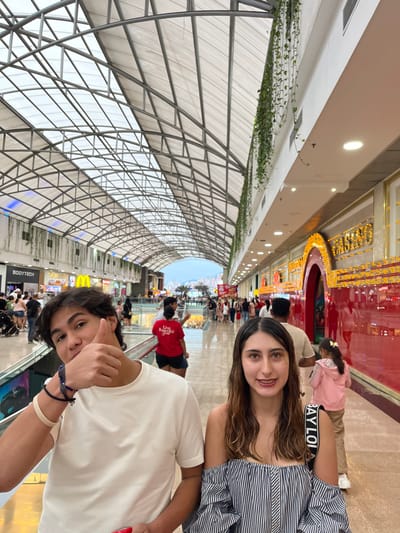 The image captures an indoor scene within Cacique El Centro Comercial in Bucaramanga, Colombia. The primary subjects are two individuals in the foreground. On the left, a male with dark, wavy hair, wearing a white short-sleeved t-shirt and multiple dark and light wristbands, looks towards the camera with his left thumb extended upwards. He appears to be holding a red object in his right hand. To his right, a female with long, wavy brown hair, dressed in a striped off-the-shoulder top and carrying a black shoulder bag, looks directly at the camera with a slight smile.
The background reveals a bustling shopping mall corridor with a distinctive arched, translucent roof structure supported by metal trusses. White walls line the corridor, adorned with vertical strands of green trailing plants. Storefronts are visible on both sides; signs for "BODYTECH" and "McDonald's" are discernible on the left. On the right, a prominent, brightly illuminated red and gold entrance sign reads "CASINO." Numerous other individuals are present in the background, walking along the corridor, including a person in a red t-shirt with "Live Beautiful" visible on the back and a young girl in a pink top near the casino entrance. The far end of the corridor opens to an exterior view of a city skyline with hills under an overcast sky, characteristic of Bucaramanga. The flooring appears to be a light-colored, reflective surface.