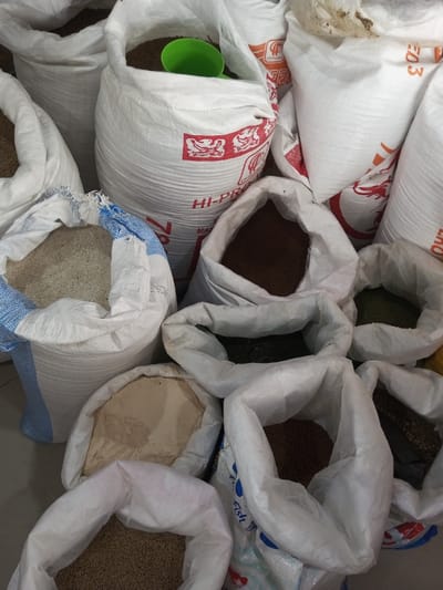 The image displays an indoor scene in Taohom, Laos, featuring numerous open white woven polypropylene bags filled with various dry bulk materials. No individuals are visible. The bags are tightly arranged on a light-colored floor or surface. Contents include light beige fine granular material, multiple types of brown granular material and pellets, dark green dried fibrous material, and light beige fine powder. A bright green plastic scoop is partially visible within one of the bags containing brown granular material. Several bags exhibit red text branding, specifically "HI-PRO" and a "CP Foods" logo featuring a crab/lobster symbol. One bag is marked with blue "Fish Food" text, and another has a distinctive blue and white striped rim. The overall arrangement indicates a retail or storage area for agricultural or animal feed products.