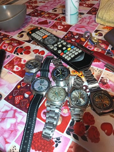 The image shows an assortment of objects on a patterned tablecloth, likely indoors. A gray bowl is visible in the upper left corner. There is a remote control, a bottle of liquid, and a towel in the upper section of the image. Several wristwatches, with metal bands and varying dial colors, are arranged together on the tablecloth. Additionally, a pair of scissors and what appears to be a knife are also present. The text from the watches and other elements are not readable, and there are no people present in the image. This image does not contain information about a city in Montevideo, Uruguay.