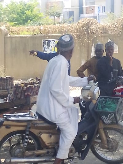 A daytime street scene in Darmanawa, Nigeria, depicts four adult males and two motorcycles. In the foreground, a male individual, dressed in a white tunic and a white and grey patterned cap, sits astride a brown and black motorcycle, his hands on the handlebars. The motorcycle features a silver exhaust pipe and a black front basket containing green fabric. To the left of this motorcycle, piles of stacked, possibly wrapped, items are present on the ground. Behind the primary motorcycle, a second male, wearing a dark blue top, extends his right arm, pointing towards the left of the frame. Further back, a tan-colored wall runs horizontally, displaying a black and yellow poster. Behind this wall, two additional males are visible: one, in a brown top and cap, stands partially obscured, while the other, in a dark top and cap, sits on a red and white motorcycle with his hand near his chin. Green trees and dried foliage are visible above the wall, with several multi-story buildings in the distant background under bright, direct sunlight.