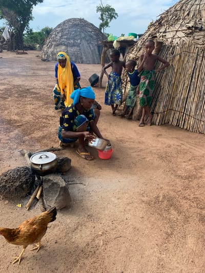 In Nandu, Nigeria, a group of individuals are gathered in a village setting. A woman wearing a blue headscarf and a patterned top is crouched down, attending to a pot cooking over an open fire made of sticks and stones. She holds a metal bowl in her hands. Next to her, another woman in a yellow headscarf and blue and yellow patterned attire stands. Three children, two girls and a boy, are also present. The children are barefoot and wear colorful patterned skirts. The boy stands slightly apart, while the girls are closer to the woman with the pot. Behind them, a conical hut constructed from thatched grass and bamboo stands, its entrance visible. Another similar structure is partially visible in the background to the left, along with trees and vegetation. A chicken is walking on the sandy ground in the foreground. The scene depicts daily life and activities within a rural Nigerian community.