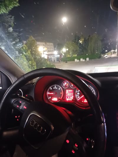 The image shows the interior of a car at night, with a view of the street and buildings through the windshield. The dashboard instruments are illuminated in red. The steering wheel is in the foreground, with the Audi logo visible. In the background, trees and buildings are illuminated by streetlights. A full moon is visible in the night sky. The scene appears to be in the city of Popovo, Bulgaria.