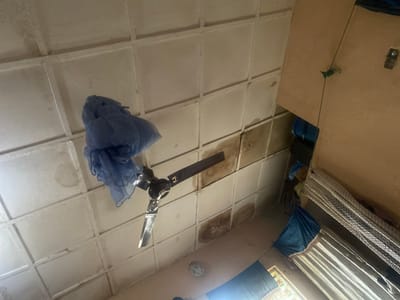 The image captures an interior view, looking upwards towards a ceiling. The ceiling is constructed of square panels, some of which show discoloration and water stains. A black ceiling fan with silver blades is mounted on the ceiling. A blue mosquito net is draped over the fan. To the right of the fan, a wall is visible, with a light fixture and a green string tied to it. Curtains, one white and one blue, hang on the right side of the image, obscuring a window. The overall impression is of a simple, possibly distressed, living space. The location is specified as Kirsinga, Nigeria.