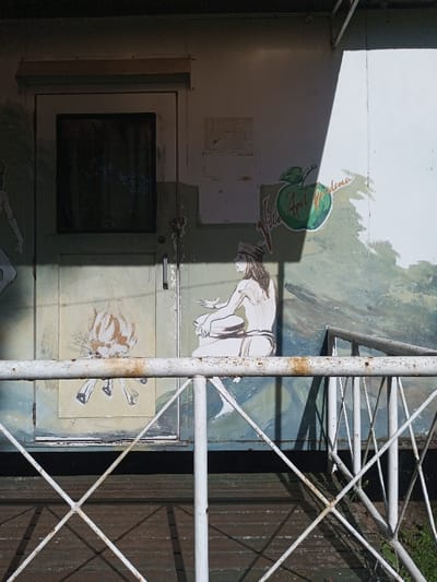 An exterior shot captures a building facade with painted designs. A door features a black-paned window and a crude fire drawing. To the right, a painted figure sits in meditation near a green apple logo reading "Vita Apfel Modena." A weathered white railing with a crisscross pattern frames the lower half of the image. The building and railing show signs of age and wear. Sunlight casts shadows, enhancing the contrast in the scene.