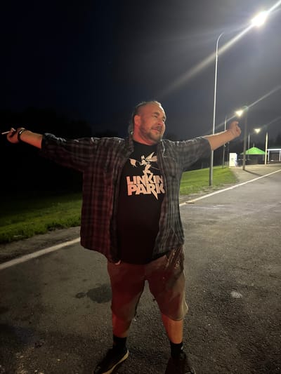 The image shows a man standing outdoors at night. He is wearing a plaid shirt over a black t-shirt with a white graphic and "LINKIN PARK" written on it. He is also wearing shorts and black sneakers with white soles. The man has his arms outstretched, and he is holding a cigarette in his left hand. He appears to be looking off to his right. The background is dark, with some streetlights visible, suggesting an urban or suburban environment. The ground is asphalt, and there is some grass and vegetation visible to the left. There is also a building with a green roof and some white structures in the background. The location is identified as Alexeyevskoye, Russia.