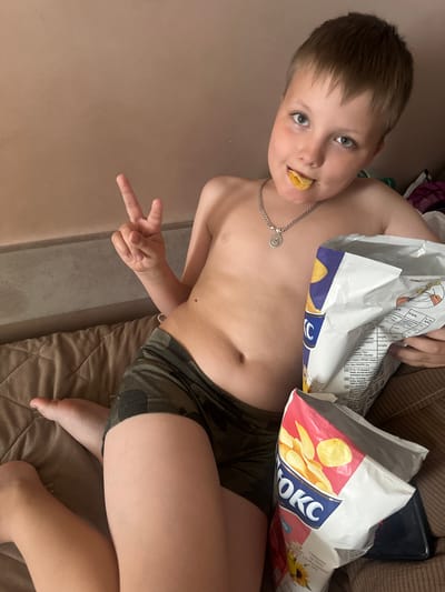 This media file shows a young boy in an indoor setting, likely a home. The boy is shirtless and wearing camouflage-patterned shorts. He is sitting on a brown couch or bed, with bags of potato chips beside him. He has a chip in his mouth and is holding up two fingers in a peace sign gesture. His expression appears playful and relaxed.
Visible text on the chip bags includes brand names and nutritional information, with text in Ukrainian and possibly other languages. Some of the text visible includes "OKC," "Смажимо на 100% соняшниковій" (Fried in 100% sunflower oil), and nutritional breakdowns like "білки" (proteins), "жири" (fats), and "вуглеводи" (carbohydrates). The location context provided is Peremyshliany, Ukraine. The lighting suggests it might be daytime or a well-lit indoor environment. The overall mood is casual and lighthearted.