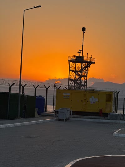 A wide-angle, eye-level outdoor shot captures a sunset over a coastal area in Gülnar, Turkey. The sky is a vibrant gradient of orange and yellow, with a few wispy clouds. In the foreground, a paved area transitions to a gravelly section. To the left, a dark green fence encloses a small structure. A tall, thin lamppost stands prominently in the left midground, silhouetted against the sky, with a bird perched on its arm.
In the midground, a chain-link fence topped with razor wire separates the foreground from the background. Behind the fence, a large yellow generator sits. To the left of the generator is a blue water tank. Further back, a dark, mountainous landscape is visible, partially obscured by clouds. A tall, metal tower structure with stairs and railings rises behind the generator. The scene is devoid of any people.