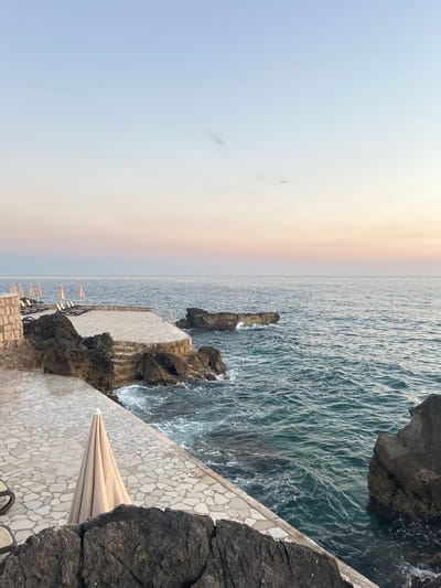 This image depicts a coastal scene in Pečurice, Montenegro. The view overlooks the Adriatic Sea, with choppy, dark blue water stretching to the horizon. The sky is a soft gradient of pale blue and peach, suggesting either dawn or dusk. Along the rocky coastline, a paved walkway with a mosaic-like stone pattern follows the shore. Beach chairs and umbrellas are set up on this walkway, indicating a resort or recreational area. Jagged rocks frame the scene, with one prominent rock formation jutting out into the sea, partially covered by the water. The overall mood is serene and tranquil, capturing a quiet moment by the sea. There is no visible text in the image.