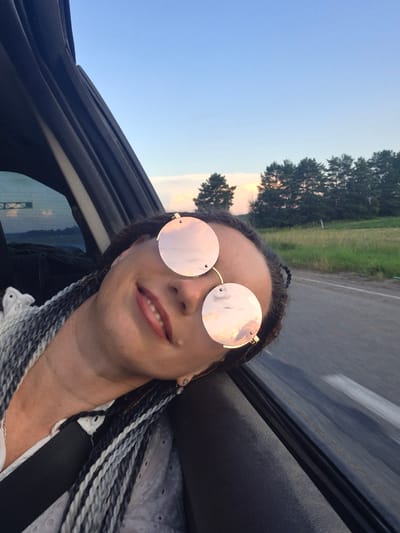 A young woman with grey and black braided hair is leaning out of a car window, looking at the camera. She is wearing large, round, rose-tinted sunglasses. The woman is smiling slightly, with her mouth open. She is wearing a white, lacy top and a black seatbelt is visible across her chest. The background shows a road, green fields, and a line of trees under a blue sky with some clouds. The image appears to have been taken in Perevoznoe, Russia.