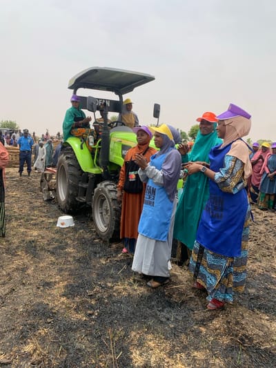 The image shows a group of people gathered around a green tractor in a field near Ringimawa, Nigeria. The tractor is parked on a field of burnt vegetation. Five women stand in front of the tractor, clapping their hands and facing the camera. They are wearing colorful clothes and head coverings. The woman on the far left is wearing a purple cap, a white shirt, and a long red and white patterned skirt. The woman next to her is wearing a blue cap, a long brown dress, and a blue apron with a white inscription. The third woman is wearing a yellow cap, a blue dress with a blue apron and a white inscription. The fourth woman is wearing a teal green head covering, and a green dress with a white inscription. The fifth woman is wearing a purple head covering, a white shirt, and a blue apron with a white inscription. Another man is sitting on the tractor, wearing a yellow cap and a blue shirt. There are several other people in the background, some wearing colorful clothes and some wearing blue shirts and white caps. The scene is bright and sunny. The field is covered in burnt vegetation. A white plastic bowl is on the ground next to the tractor. The location of this image is coordinates (latitude: 11.666, longitude: 8.416) near Ringimawa, Nigeria.