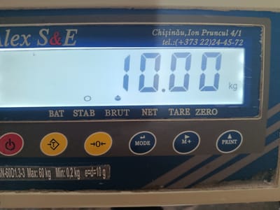 The image shows a close-up of a digital scale. The scale displays a weight of "10.00" and "kg". Above the display, text reads "Alex S&E" and "Chisinau, Ion Pruncul 4/1 tel.:(+373 22)24-45-72". Below the display, there is a row of buttons, including "POWER", "TARE", "MODE", and "PRINT". There is also a label that reads "SN-60D1.3-3 Max: 60 kg Min: 0,2 kg e=d=10 g". The scale appears to be situated indoors, with a blue and grey casing. The lighting is bright and somewhat uneven. There is no indication of the location being in the city of Bălți, Moldova; the address provided is in Chișinău.