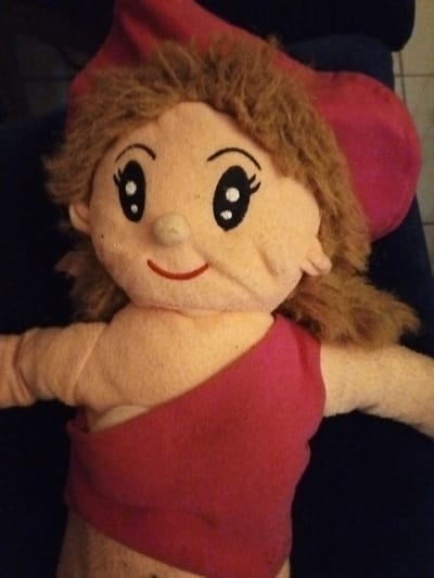 A fabric doll with light skin, brown yarn hair, and a red hat lies against a dark blue surface. The doll has cartoonish eyes with eyelashes and a small red smile. She wears a red fabric garment draped across her chest. The doll’s stuffing appears slightly uneven, particularly around the face and torso, and shows some wear.