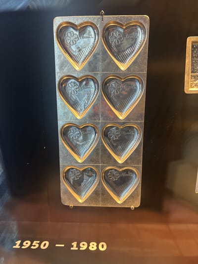 A metal mold containing eight heart-shaped cavities sits in a display case. Each cavity has a slightly raised outline and depicts a four-leaf clover and the word "Glück" in script. The display case is black, and to the right, on a dark brown panel, the years "1950 - 1980" are printed in bold, off-white letters. The location is likely a museum in Kilchberg (ZH), Switzerland, showcasing items from that time period, but no humans, actions, or interactions are visible in the frame.