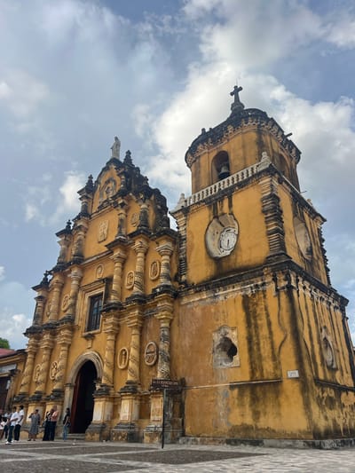 The image shows the yellow facade of the Iglesia de la Recolección in León, Nicaragua, under a cloudy sky. The church features intricate architectural details with columns, sculptures, and a prominent bell tower with a clock. A statue of a religious figure stands atop the main building. Several people are gathered in front of the church entrance, and a sign indicates the church's name and construction year of 1786. The ground in front of the church is paved with stone tiles.