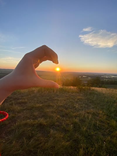The image captures a sunset over a rural landscape, likely in or near Уязытамак, Russia. A human hand is positioned in the foreground, forming a heart shape around the setting sun. The sun is a bright, golden orb, casting long rays across the sky and illuminating the scene with warm light. The terrain is characterized by rolling hills covered in dry grass, indicative of a summer or early autumn setting. In the distance, a patchwork of fields and bodies of water can be seen, suggesting a sparsely populated area with agricultural activity. The sky is a gradient of blue and orange, with a few wispy clouds adding texture. A red and yellow rope object is partially visible in the lower left corner of the frame.