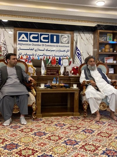 This medium shot shows two men in an office. The man on the left wears a gray suit and light gray socks. The man on the right wears a white outfit with a white head covering. The men are sitting in ornate chairs on opposite sides of a table. On the table are water bottles and two cups.

Behind the table is the Afghanistan Chamber of Commerce & Investment (ACCI) logo and several small flags of different nations. In the background on the right, there is a shelf with decorative items. The carpet is patterned.

There is no other information to indicate the location as Arghandab, Afghanistan.