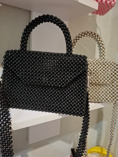 The image shows a close-up view of two beaded handbags displayed on white shelves. The foreground handbag is black and made of small, shiny black beads. It has a top handle and a shoulder strap, both also constructed from beads. The background handbag is a lighter color, possibly silver or pearl, and is made of similarly sized beads. It also features a beaded handle and strap. In the upper right corner, a portion of a pink beaded bag is visible. The lighting is soft, highlighting the texture and shine of the beads. The environment appears to be an indoor display, likely a retail setting. The image was taken in Korenovsk, Russia.