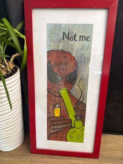 The image shows a framed drawing. The drawing depicts Spider-Man holding a bong and a lighter. The text "Not me" is written above Spider-Man's head. The frame is red and the matting is white. The drawing is in a vertical orientation. The drawing is placed on a wooden surface next to a white potted plant. The background is dark. There is no information available about the location in the city of Хохряки, Russia.