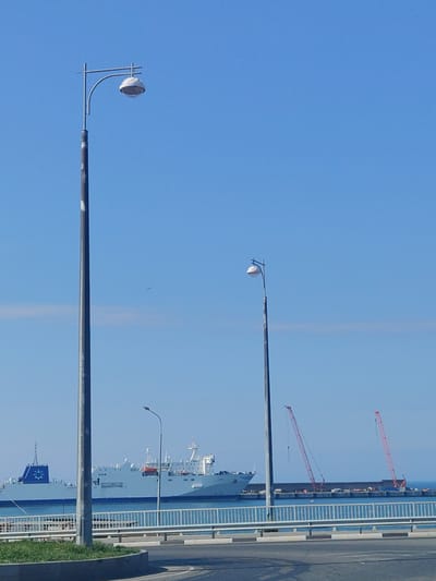 The photograph depicts a sunny, clear day in Pionersky, Russia, featuring two tall, grey lampposts in the foreground. Behind the lampposts, a grey passenger ship is visible on the water, and a pier with two red cranes. A white metal railing separates the road from the sea. The sky is a clear blue.