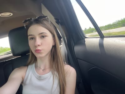 A young woman with long brown hair is sitting in the passenger seat of a car. She is wearing a white ribbed tank top and a silver heart necklace. Her sunglasses are perched on top of her head. The interior of the car is visible, including the black leather seats and the car's ceiling. Outside the car window, a road and lush green trees can be seen. The setting appears to be a rural or suburban area.