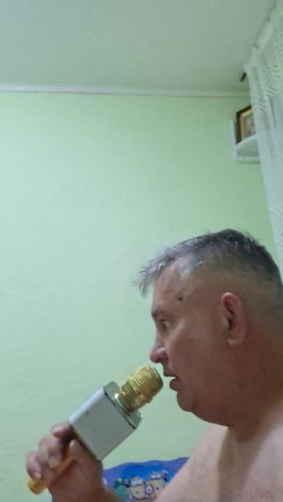 An adult male with short, graying hair and a mole on his left cheek is holding a white and gold microphone to his mouth. He is seen in profile, facing right, with his mouth slightly open as if singing or speaking into the microphone. The background consists of a pale green wall and a white curtain on the right side of the frame. A small framed picture is visible on a shelf in the upper right corner. The man appears to be indoors, in what could be a residential setting in Voinova, Moldova, based on the available context.