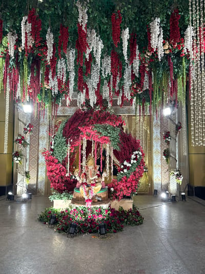 An elaborately decorated indoor or covered outdoor venue in Bidhannagar, India, is depicted. The scene features a central floral archway framing a multi-colored statue of Lord Ganesha seated on a pedestal. The archway is densely arranged with red, pink, white, and green artificial flowers and foliage. The Ganesha statue is light-toned with a yellow garment and appears to hold various items in its four arms, adorned with a small floral garland. The base surrounding the statue is covered in dark pink flowers and green foliage.

Overhead, the ceiling is blanketed with green foliage, from which numerous strands of red, white, and green artificial flowers and white bead-like strings hang vertically. The background consists of ornate wall panels with repeating patterns and a grand doorway. Additional floral arrangements featuring red and white flowers are mounted on the side walls. Multiple black spotlights are positioned on the light gray floor, illuminating the floral display from below. Square white floodlights are also visible within the overhead decorations. No individuals are present in the image.