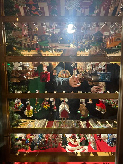 A display case holds various Christmas ornaments, including wooden figures, houses, and trees. Garland decorates the bottom. The window reflects a person with glasses and light flares across the image. The ornaments are hung on wooden dowels with price tags attached. Red and green are prominent colors among the decorations. The case has a wooden frame with vertical bars.