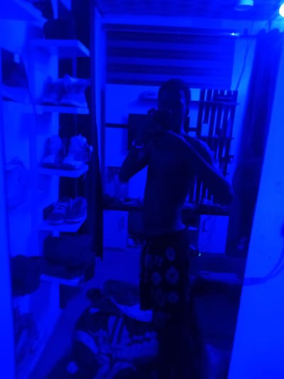 The image captures a person taking a selfie in a room illuminated by blue light. The person is wearing dark shorts with a floral pattern and is shirtless. Behind them, shelves are visible, holding several pairs of sneakers. To the right, a mirror reflects the scene. The overall environment appears to be a domestic setting, likely a bedroom or closet, in the city of Ogbogbo, Nigeria. The lighting creates a dramatic and moody atmosphere.