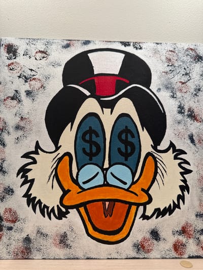 The image displays a close-up, eye-level view of a painted canvas featuring a cartoon duck character with dollar signs in its eyes. The duck is wearing a black and white striped hat with a red band. The background of the canvas has a textured, splattered appearance with flecks of black, red, and blue paint on a white base. The painting is positioned against a plain white wall, and the bottom edge of the canvas rests on a light-colored wooden surface. There is no discernible information within the image to indicate a specific location in Biržai, Lithuania.