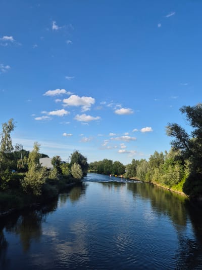 This is a wide-angle, vertical shot of the Isar River in Landshut, Germany, on a bright, sunny day. The sky is a vibrant blue, dotted with fluffy white cumulus clouds. The river flows from the foreground towards the background, its surface rippling and reflecting the sky and surrounding foliage.
The riverbanks are lined with lush green trees and bushes, creating a natural, verdant landscape. On the left bank, a white building is partially visible amidst the greenery, and a car can be seen parked to its left. Further in the distance on the left bank, tall trees with distinctive, slender trunks are prominent. On the right bank, the trees are denser and form a more continuous line of vegetation.
In the middle of the river, slightly upstream, there are two navigational markers. One appears to be a yellow diamond shape, and the other is a red and white striped marker. The water seems to flow faster around these markers, indicating a slight change in the riverbed or current. The overall scene depicts a serene and natural environment within the city.