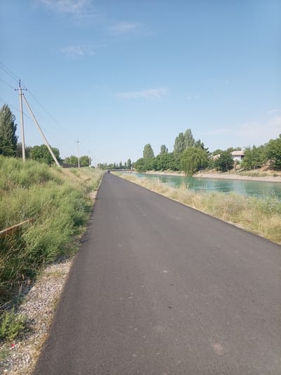 A paved road runs alongside a wide, turquoise canal in Azadbash, Uzbekistan. The road is empty except for a car in the distance. On the left side of the road, a grassy embankment slopes upwards, dotted with sparse trees and utility poles. The right side of the canal is lined with trees and a few houses. The sky is clear and blue with a few wispy clouds. The scene appears serene and rural.